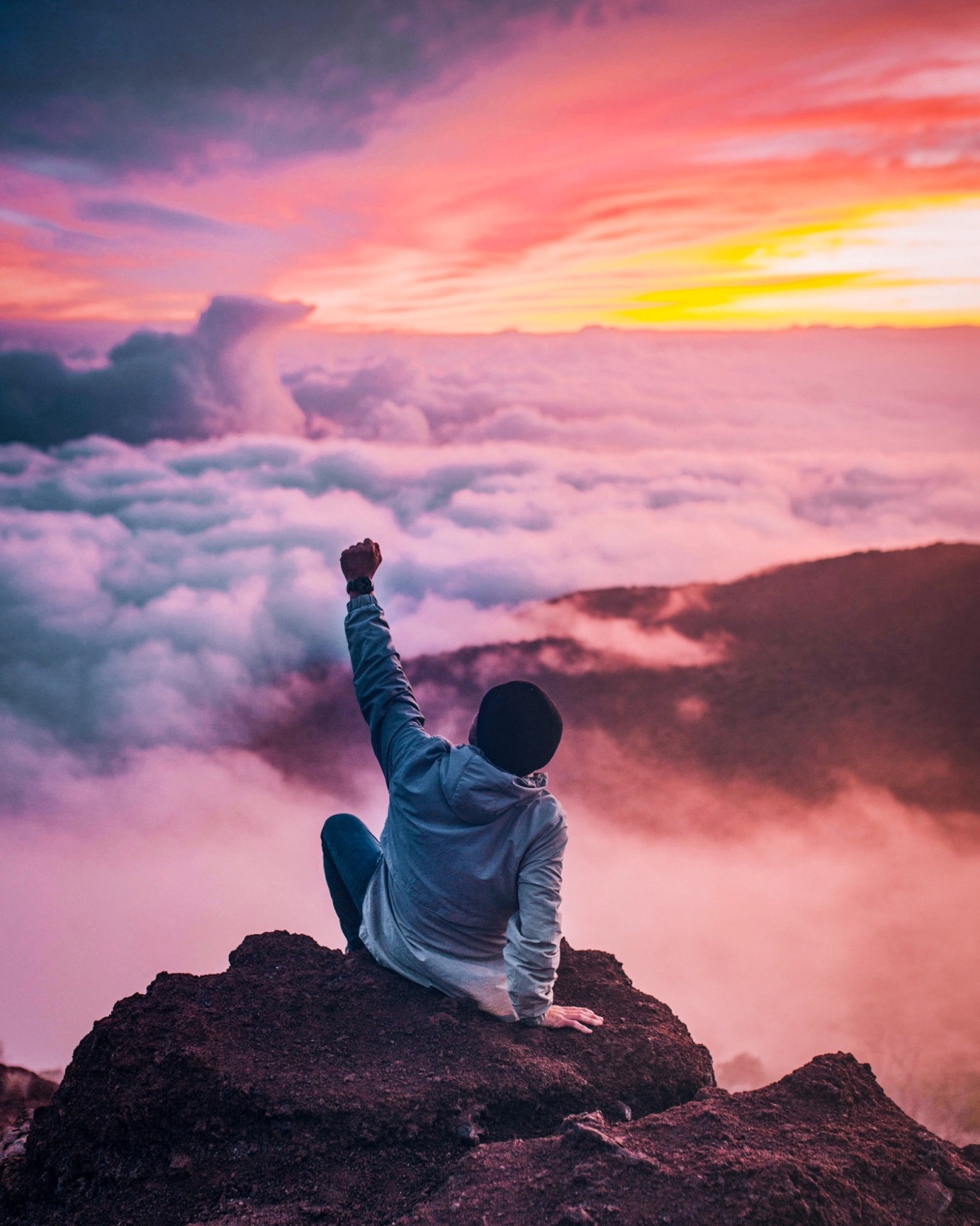 A person sitting on a cliff, facing a sunset and having a fist up for showing success.